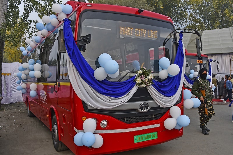 A person holding a National Common Mobility Card (NCMC) and swiping it on a payment terminal in an e-bus, with a blurred background of Srinagar cityscape. The NCMC logo and SSCL logo are prominently displayed. The image represents the launch of NCMC service by Srinagar Smart City Limited (SSCL) for convenient cashless travel in e-buses.